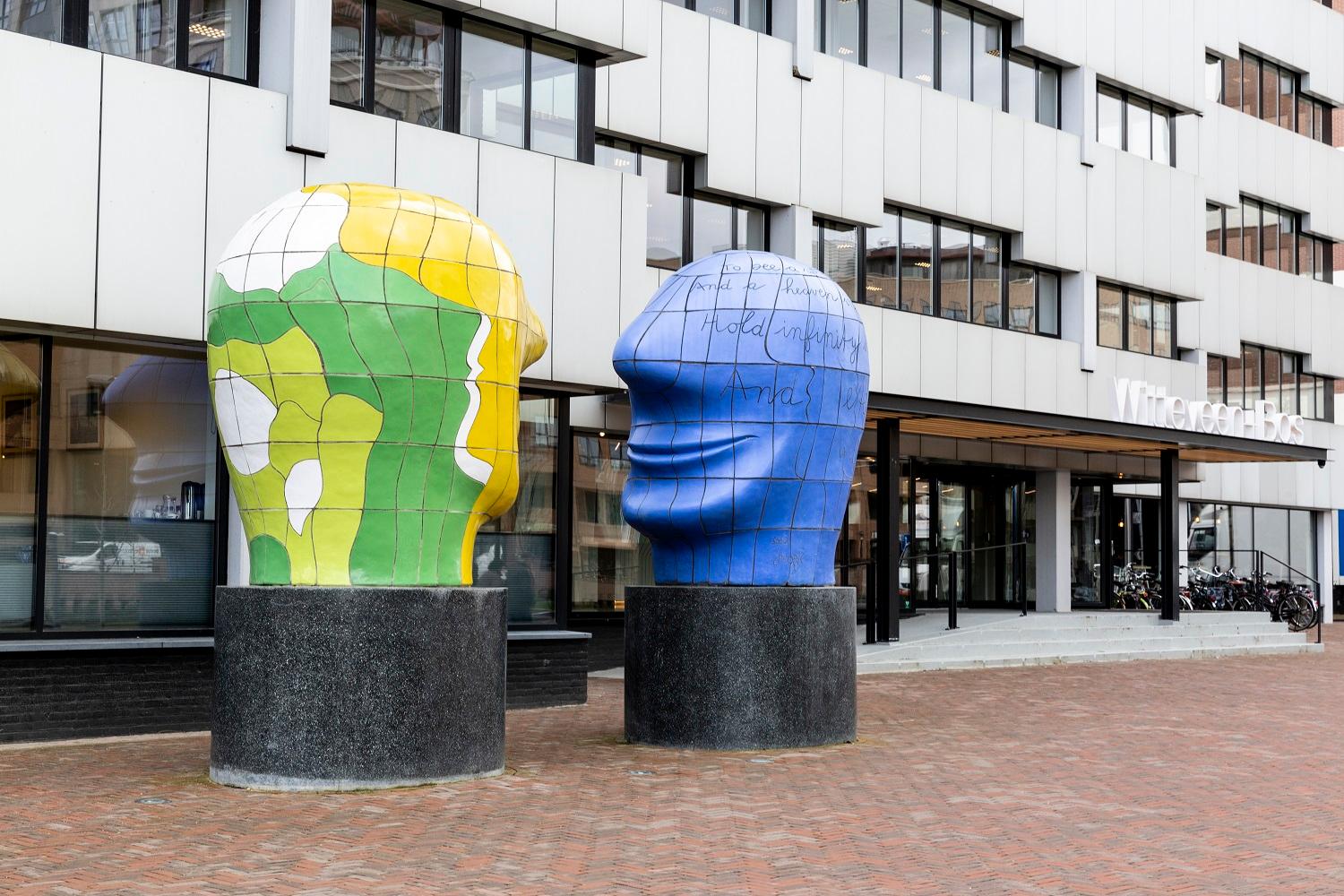 Zij aanzicht van het hoofdkantoor in Deventer. Het pand is wit met zwarte ramen en er staat een kunstwerk voor van twee grote hoofden.