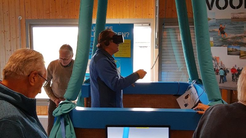 Er staat een man met een VR-Bril in de mand van de luchtballon op de grond. Op een televisiescherm zie je hoe de man boven de dijk vliegt.