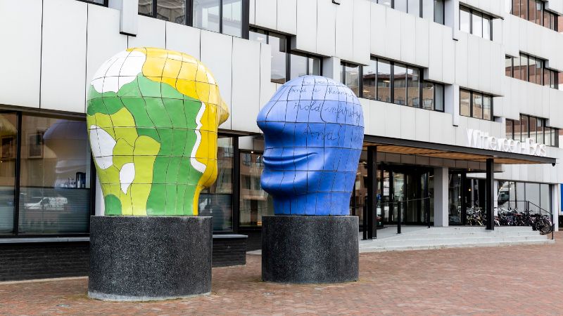 Side view of the headquarters in Deventer. The building is white with black windows and there is an artwork of two large heads in front of it.<br/>
