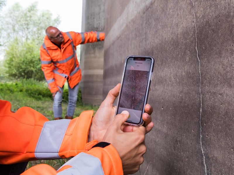 Twee handen houden een telefoon bij een scheur in de muur. Je ziet de Crack Width Analysis-app, die de muur scant. Op de achtergrond staat nog iemand de muur te controleren.