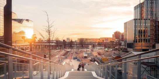 A photograph of a bridge with 2 people walking on it.  The sun is reflected in the buildings.