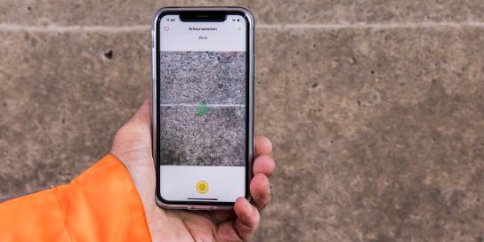 A phone near a crack in the wall. On the screen is the Crack Width Analysis app.