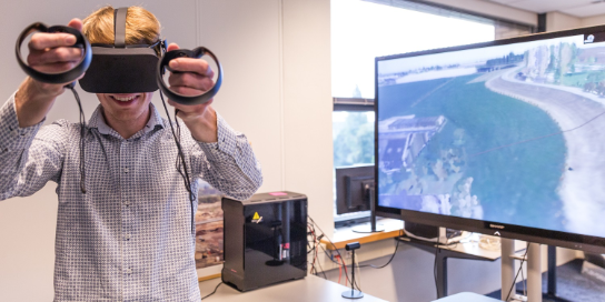 A boy is wearing VR glasses. Next to him is a screen showing how he is flying above the dyke.