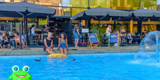A summery photo of two young women sitting with their legs in a basin of water to cool down. In the background is a terrace.