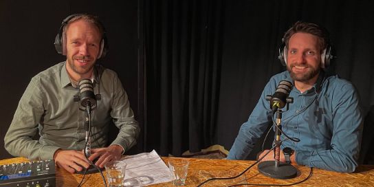 A photo of Rony Nedkov and Pieter-Bas de Visser. They are sitting behind a microphone and wearing both headphones.