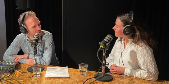 A photo of Eva Geerlings and Pieter-Bas de Visser. They are wearing headphones and are sitting behind a microphone.