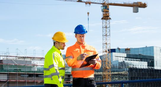 Two men wearing helmets and yellow vests are looking at a memo. They are standing on a building site. In the background is a large crane.