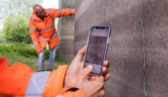 Two hands are holding a phone near a crack in the wall. You can see the Crack Width Analysis app, which is scanning the wall. In the background, another person is checking the wall.