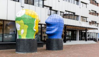 Side view of the headquarters in Deventer. The building is white with black windows and there is an artwork of two large heads in front of it.<br/>